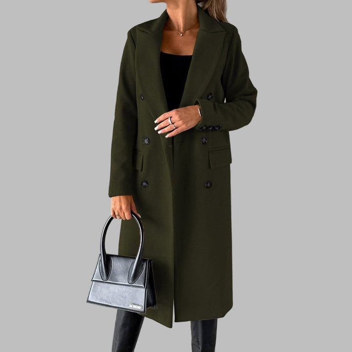 Vrouw in olijfgroene winterjas met dubbele knopen, zwarte handtas en zwarte laarzen tegen een grijze achtergrond. Stijlvolle damesmode.